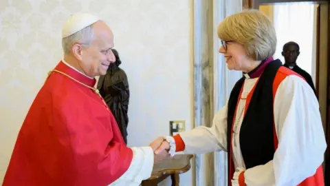 Vatican Media/PA Pope Leo XIV, a smiling white man with grey hair, wearing a red robe and a white skull cap, shakes the hand of Dame Sarah Mullally. She is wearing white, red and black religious robes with a large cross around her neck and a clerical collar. Her hair is blond and short, with a fringe and she is smiling.
