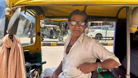 BBC Mohammad Umar sits in his autorickshaw. He is wearing a white shirt and white trousers and wearing smudgy glasses.
