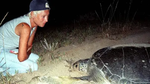AFP via Getty Images Mona Khalil, wearing a navy blue cap backwards, looks at a turtle on the coast in the southern Lebanese port city of Tyre in 2002.