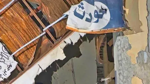 WHO Ruins of walls and a ceiling at el-Daein Teaching Hospital. A blue and white sign with the word reading 'Accidents' in Arabic is seen hanging in tatters.