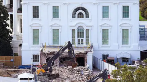 Getty Images The side view of the White House that shows a wrecker machine out front, on a large pile of rubble. In the foreground there is other machinery for demotion and some green trees. 