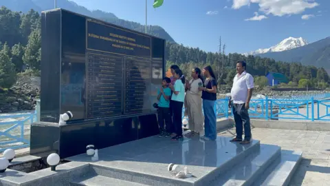 Tourists in a group of six - four children and their parents - stand in front of the memorial built in Pahalgam for the 26 people who were killed by militants here last year.