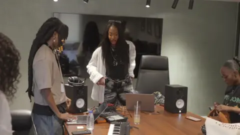Tems looks at a young woman, who is using a laptop connected to various musical equipement