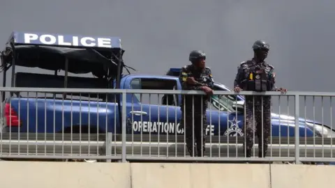 Anadolu Agency via Getty Images Two police officers leaning over a white railing outdoors. Behind them is a blue police vehicle and a police tent