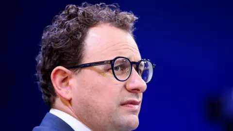 Reuters Anthropic CEO Dario Amodei on stage wearing a blue blazer and dark-rimmed round eye glasses.