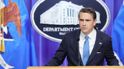 Getty Images Todd Blanche wears a blue tie and suit. He stands in front of a Department of Justice backdrop as he speaks into a microphone