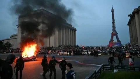 AFP via Getty Images Police officers near a fan in the area near Eiffel Tower which can be seen in the distance with a large number of people gathered; a car is on fire in the middle of the road, in from of a tall building