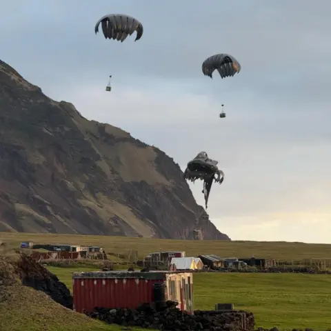 Minstry of Defence Parachutes delivering boxes of military supplies seen against a rugged backdrop, where several small buildings can be seen 