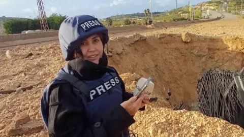 AP Photo of Al Akhbar journalist Amal Khalil, wearing a press jacket and helmet, reporting near a destroyed bridge in Qasmiyeh.