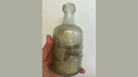 Deb Brown via AP A hand holds an old green glass bottle with paper inside it against a wall