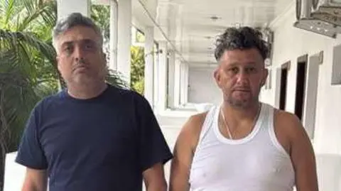 Courtesy image Carlos Alberto Rodelo, in blue T-shirt and shorts, stands next to Jorge Cubillos, in white vest and black trousers, with hands in pockets, on the balcony of their small hotel room in Kinshasa, with palm trees in the background.