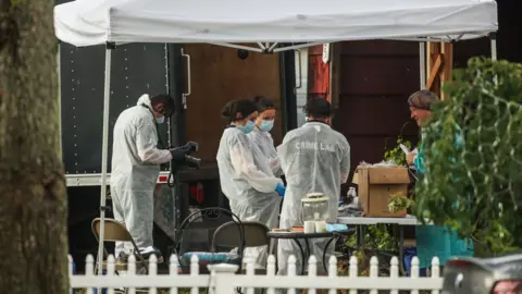 Getty Images Investigators in white coats, hairnets and jackets labelled "crime lab" inspect items outside a home under a white tent