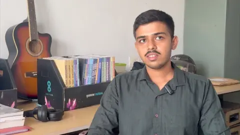 Zoya Mateen Arman dressed in a black shirt, sits in his hostel room. There are books and a guitar lying behind him on the table 