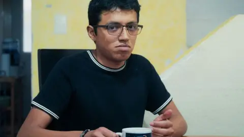 Julian is wearing a navy and white t-shirt and is holding a cup of tea - he has glasses and dark hair