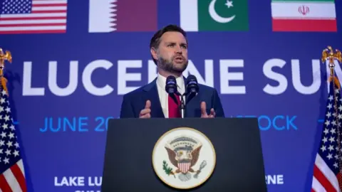 Reuters JD Vance speaking at a podium - he is holding his hands to make a gesture while speaking and looking out. He is flanked on either side by American flags, with the flags of Iran, Qatar, Saudi Arabia and the US visible on a graphic behind him. 