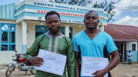 Reuters Tagirabo is wearing green and Bulambula is wearing turquoise. they are each holding a white certificate they received for recovering, and are standing outside a medical centre