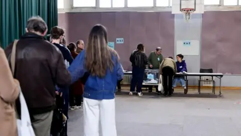 EPA Voters queue inside a polling station at the far end of which two officials at a long table face two of the voters about to retrieve their ballots