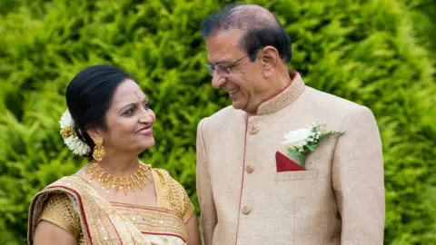 Family handout A man and a women stand outdoors against a backdrop of green leaves, wearing detailed traditional outfits. They are standing close together and looking away from the camera and at each other, smiling.
