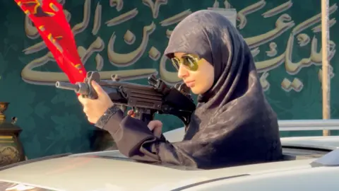 A young woman in a headscarf and sunglasses is seen standing up through the sunroof of a car, holding a large gun, as part of a pro-regime demonstration.