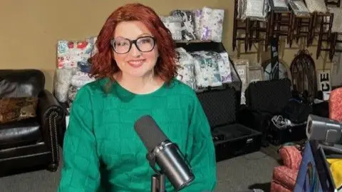 Anthony Zurcher/BBC Tiffany Ciani, an influencer who supports Thomas Massie, sits in front of a microphone and laptop in a room filled with furniture for sale. 