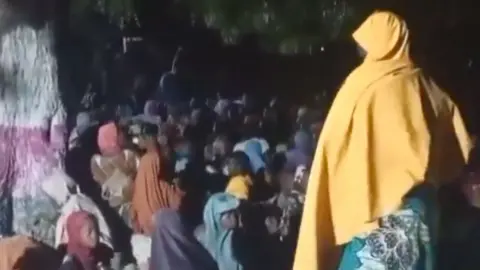 @HQNigerianArmy A screengrab from a video showing a crowd of mainly and children, mostly from behind, as they sit under a treat after being freed from Boko Haram captivity. One of the women is standing to the right of the image and wears a yellow headscarf.