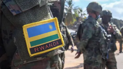 AFP via Getty Images A close-up of a soldier’s uniform showing a Rwandan flag patch on the shoulder. Armed soldiers stand in the background.