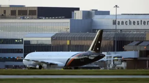 PA Media An airplane is on the runway, with grass in the foreground and an airport in the background with Manchester emblazoned on it