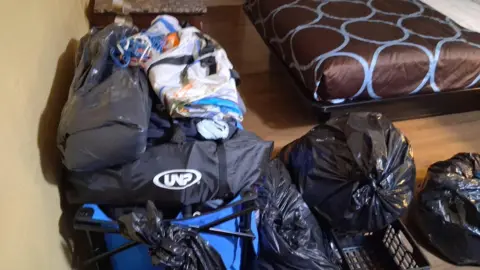 Submitted photo Duffel bags and full garbage bags are piled up on a floor next to a bed with a brown duvet