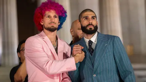 Anadolu via Getty Images Two fighters - one with pink and blue hair wearing a pink suit and another wearing a pinstripe suit - bumping fists in front of the Lincoln Memorial