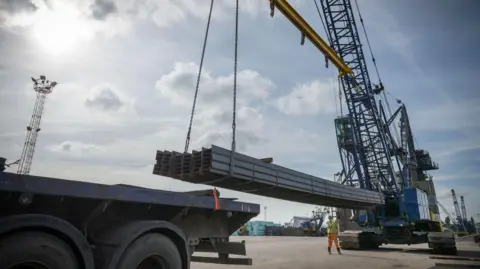 BBC Large steel rods are lifted with a crane. It is a sunny day with blue skies. 