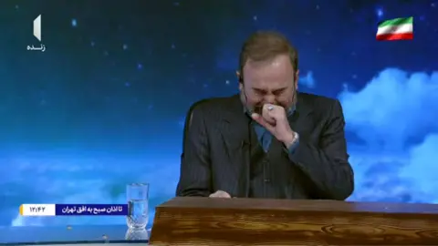 IRTV Image shows a presenter on TV wearing a dark suit, with one hand to his face, eyes closed, and crying. The background behind him shows a blue sky with clouds