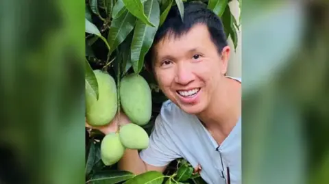 ABC Thanh Phan, looking at camera, wearing white t-shirt, posing with a mango tree with four green mangoes on the branches