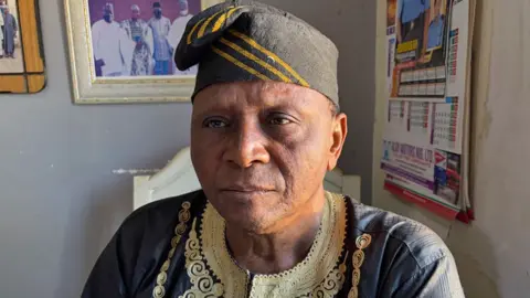 BBC/Alex Last His Royal Highness Chief Dr Danladi Akinga Kasuwa sitting in an office with a calendar and framed photographs on the walls. He is dressed in a traditional dark near-black robe, with a yellow embroidered design around the neckline, and a near-black woven hat decorated with three mustard coloured stripes.