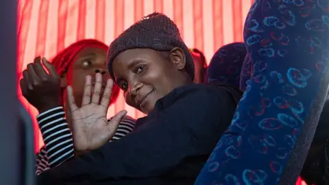 AFP via Getty Images A woman in a grey hat sitting in a coach smiles and waves from the window at the people outside. 