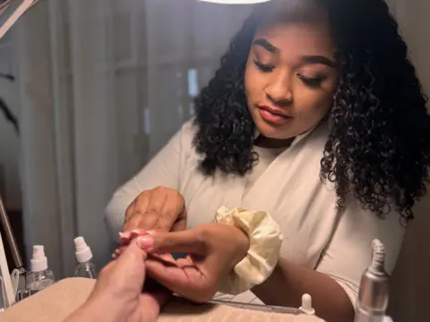 Close-up of hands during a manicure, with one hand held over a beige towel while another applies product to the fingernails. Small bottles with pump dispensers and a dropper sit on a tabletop in the foreground. In the midground, a person with curly hair leans over the hands under a bright desk lamp, wearing a light-coloured top and a satin scrunchie on the wrist. The background shows a softly blurred interior with neutral tones and indistinct furnishings.