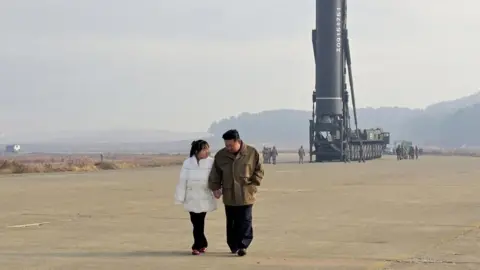 Reuters With her long hair tied back, in black trousers and a white padded jacket, Kim Ju Ae walks hand-in-hand with Kim Jong Un, in a brown jacket and black trousers. They are looking towards each other. Towering above them, with several men standing next to it, and hills in the background, an intercontinental ballistic missile (ICBM) points upwards, on a caterpillar-track transporter.