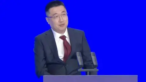 Screenshot from Guangdong News Shein founder Xu Yangtian, or Sky Xu, speaks to an audience during the Provincial High-quality Development Conference. He is standing behind a lectern while dressed in a navy suit and burgundy tie. 
