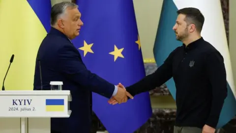 Reuters Orbán wearing a dark suit shakes hands with Zelensky who is wearing a black top and green trousers - with the European Union and Hungarian flags in the background and a white podium wiehere Kyiv and 2 July 2024 is written alongside the Ukrainian flag colours.