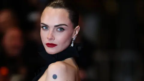 Getty Images Cara Delevingne has dark hair scraped back into an up-do. She has red lipstick on and is wearing a dress with a black halterneck and open back. 