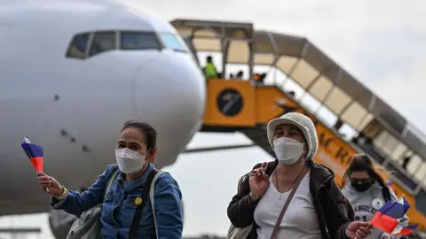 Getty Images Three women waving the Philippine flag and wearing face masks, walking from an airplane in the background after being repatriated fro the Middle East due to the US and Israel's war on Iran