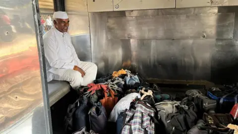 Shahid Sheikh A man wearing a white kurta pajama and a cap sits in a train berth with lunchboxes placed on the floor