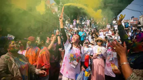 Getty Images KATHMANDU, NEPAL - 2026/03/02: A woman smeared with vermilion powder fires a color smoke bomb during the Holi festival celebration. The Holi festival is celebrated to mark the victory of good over evil and also symbolizes the arrival of the spring season.