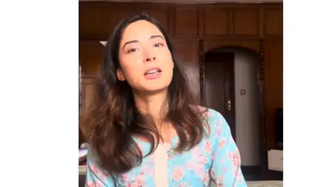 Shrinkhala Khatiwada/YouTube Shrinkhala Khatiwada sits in a room with cupboards in the background. She is wearing a blue top with pink flowers on it. It is a video shot in vertical and on a YouTube channel