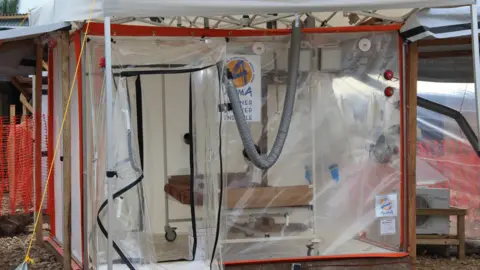 Jennifer Lazuta/ALIMA A plastic, transparent cube-like structure with a zip-up door that is open. There is an empty bed inside and no patient