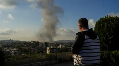 Reuters A man in a grey and black striped jacket looks at smoke rising from a building following an Israeli airstrike, he is holding a smart phone.