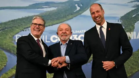 PA From left to right, Sir Keir Starmer, President Lula and Prince William. All are wearing dark-coloured suits. Sir Keir is wearing a white shirt and dark red tie; President Lula a light blue shirt; and Prince William a white shirt and blue tie. They are putting their hands together in an apparent show of unity. Blue and green COP30 banners are in the background.