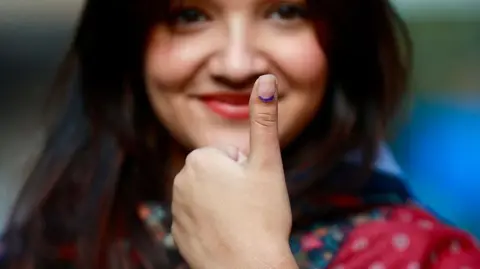 AFP via Getty Images A female voter with long dark hair and wearing a red patterned dress shows her inked thumb after casting her ballot at a polling station during Nepal's parliamentary election in Kathmandu on March 5, 2026
