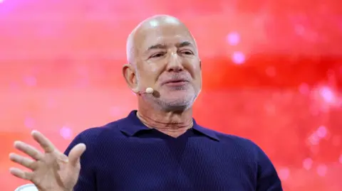 Reuters Jeff Bezos wearing a headset microphone and a navy blue polo shirt. He is on stage in front of a bright red background