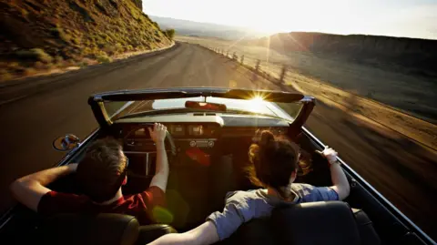 Getty Images A young man and woman in a convertible car driving along a mountain road towards the sunset. She is driving and has her arm around the back of his seat.