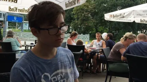David Parfett Thomas Parfett, a young man wearing a faded blue T-shirt and black-framed glasses. He is sat in an outdoor seating area with parasols.